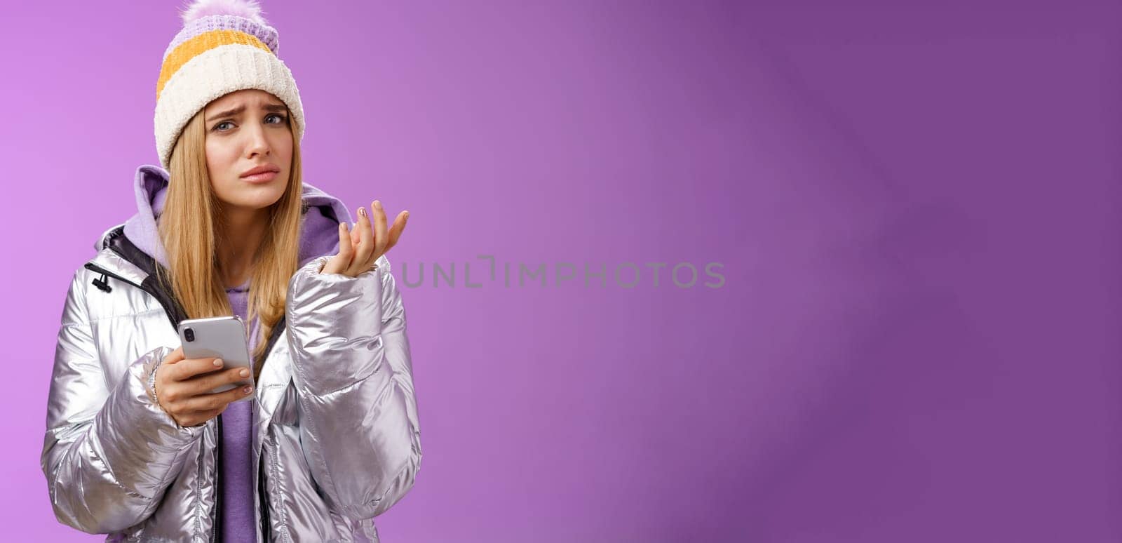 Questioned complicated cute blond girlfriend receive strange message look perplexed confused raising hand shrugging lift eyebrow cannot understand meaning holding smartphone, purple background.