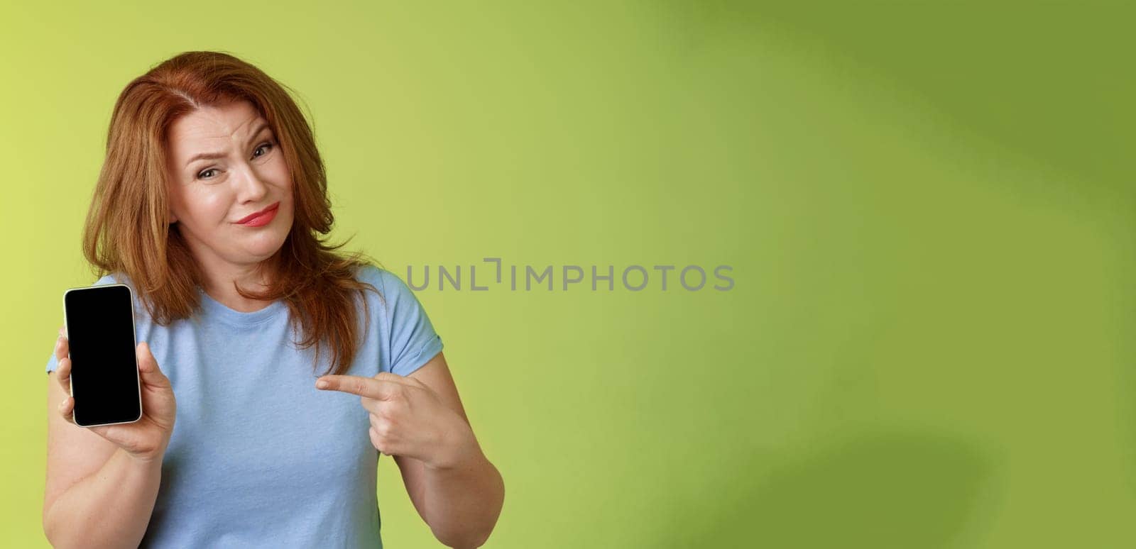 Seriously it awful. Displeased disappointed redhead mature female tilt head cringe grimacing reluctant pointing smartphone blank display index finger showing bad photograph share negative opinion.