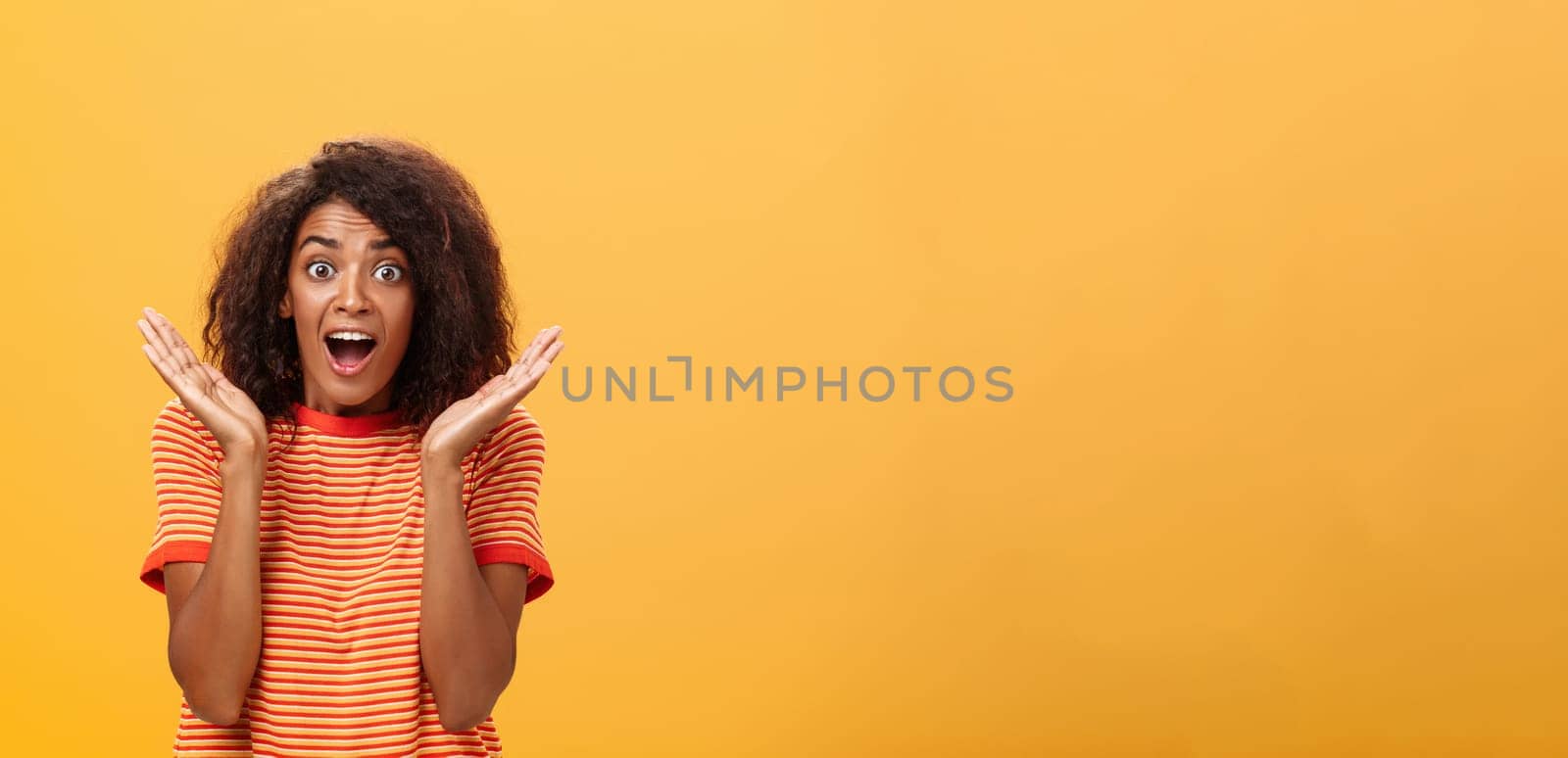 Waist-up shot of amazed joyful sociable dark-skinned female with afro hairstyle in striped t-shirt gesturing with palms raised near face gazing astonished and delighted at camera from surprise and joy. Lifestyle.