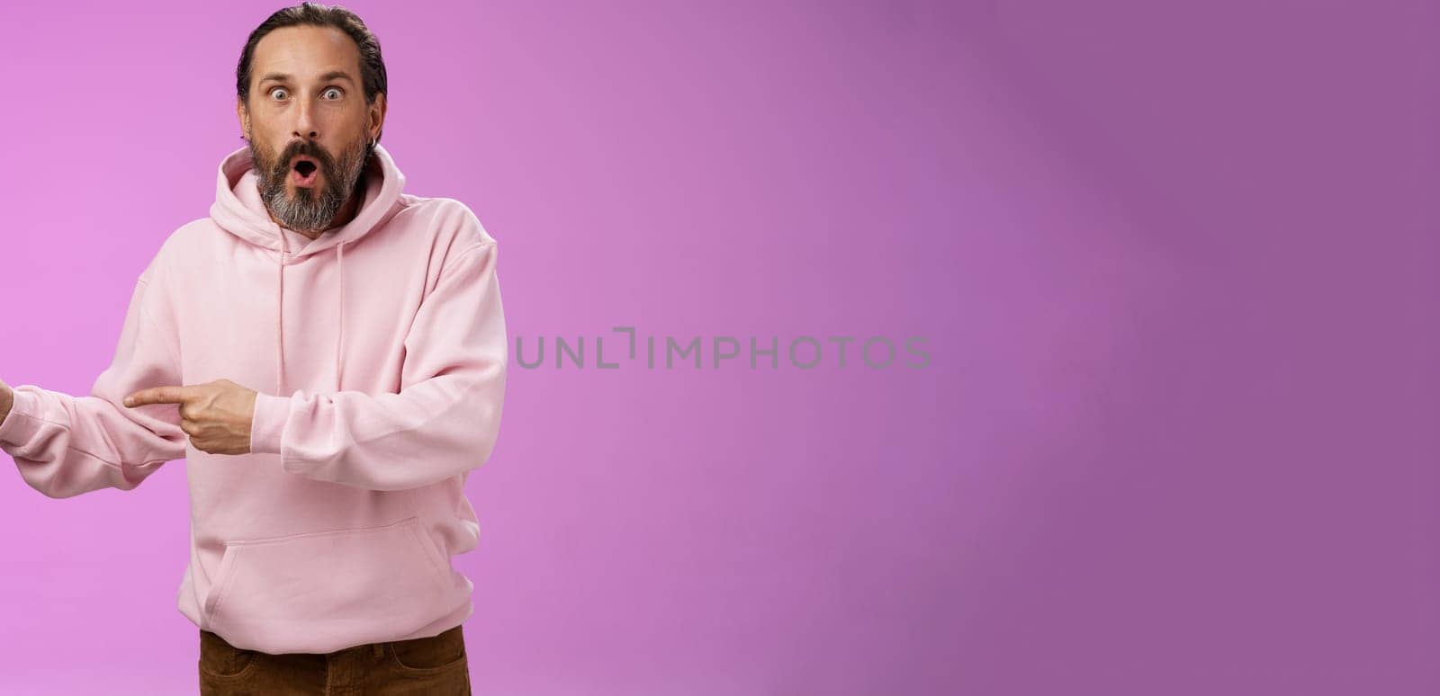 Astonished fascinated curious mature bearded man with grey hair in pink hoodie folding lips amazed pointing left index fingers widen eyes camera impressed thrilled wanna go check out cool promo. Lifestyle.