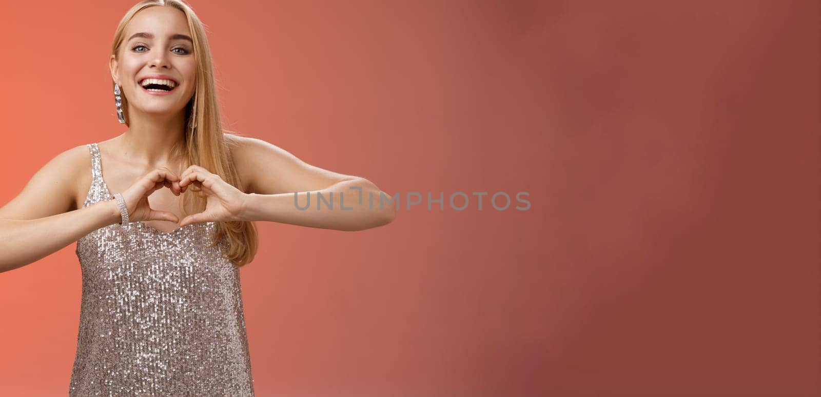 Adorable happy charming lovely blond european girl in silver dress express love positivity show heart gesture confess sympathy passionate feelings towards boyfriend smiling broadly, red background.