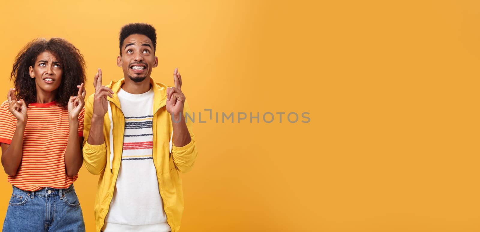 Couple of friends praying and looking up in sky intense crossing fingers for good luck clenching teeth faithfully hoping parents not saw them smoking standing together over orange background by Benzoix