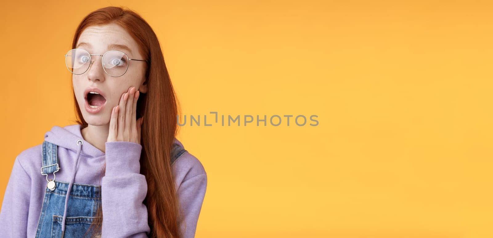 Shocked impressed speechless young redhead sensitive european girl glasses wearing hoodie dungarees drop jaw gasping astonished look left wide eyes amazed standing orange background.