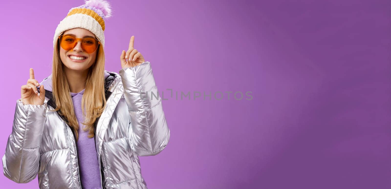 Lifestyle. Joyful energized entertained cute blond woman having fun enjoy vacation snowy mountain trip wearing sunglasses silver jacket winter hat dancing pointing up amused standing purple background.