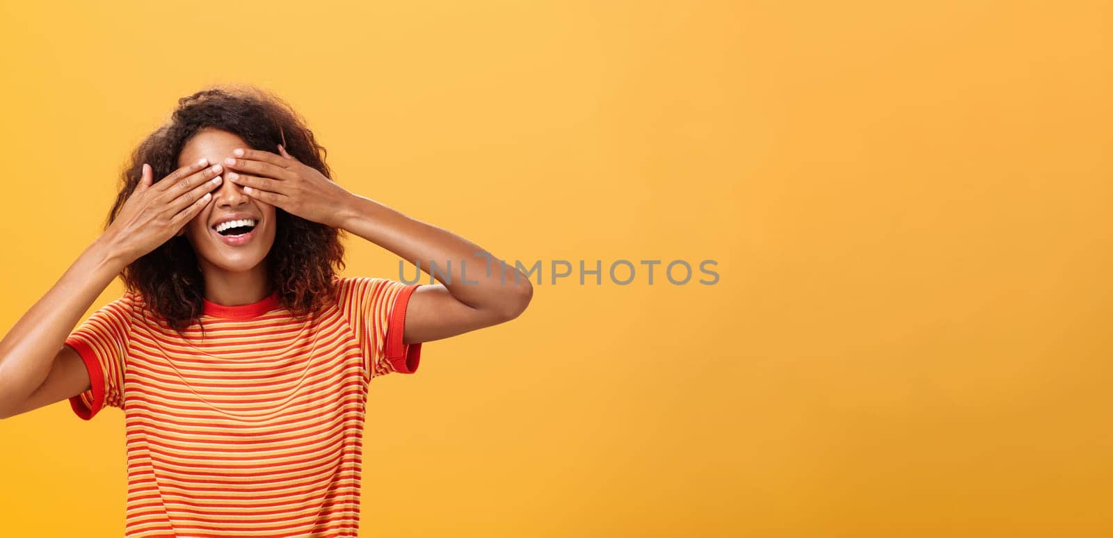 Closing my eyes and counting ten. Portrait of charming dreamy and happy funny african american curly-haired female in striped trendy t-shirt covering sight with palm and smiling waiting surprise. Copy space