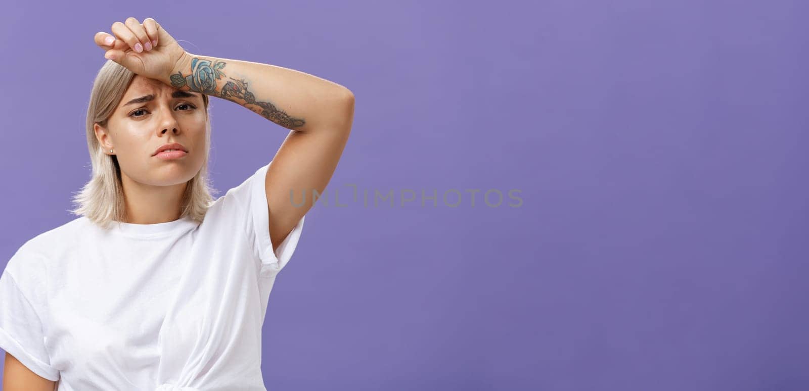 Lifestyle. Indoor shot of tired and displeased exhausted cute european woman with tanned skin and tattoos on arms holding hand on forehead frowning suffering from tiresome work or sun heating over purple wall..