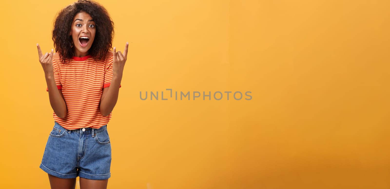 Stylish african american woman being delighted and excited before cool concert showing rock n roll gesture with both hands screaming from satisfaction and happiness cheering over orange background. Copy space