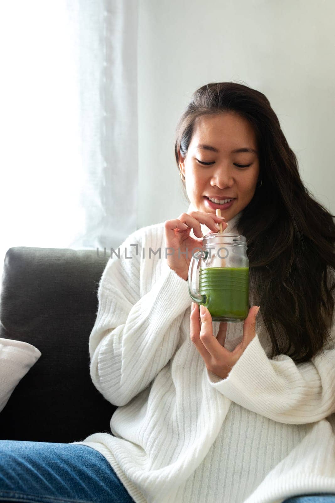Vertical portrait of young beautiful Asian woman relaxing at home drinking healthy green juice. Female enjoying vegetable smoothie. Healthy eating and lifestyle concepts.