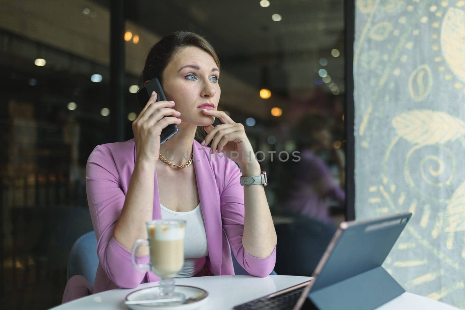 Thoughtful business woman talking on the phone with clients or business partners, making difficult decisions while sitting in coffee shop with latte and laptop.