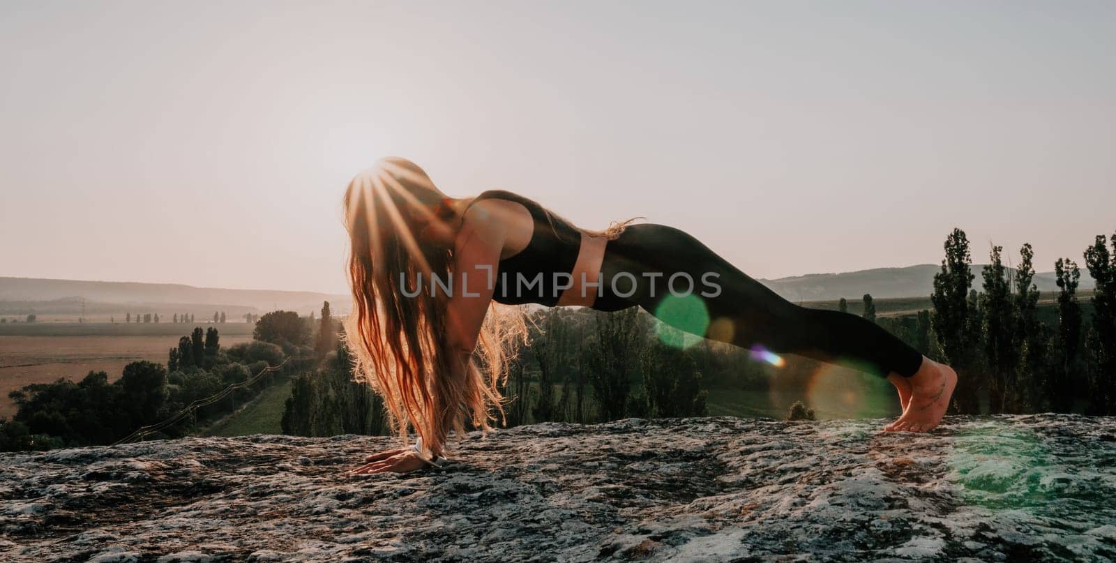 Well looking middle aged woman with long hair, fitness instructor in leggings and tops doing stretching and pilates on the rock near forest. Female fitness yoga routine concept. Healthy lifestyle.