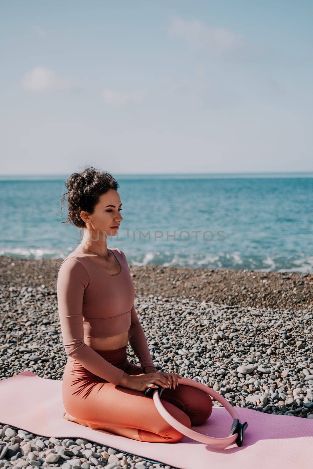 Middle aged well looking woman with black hair doing Pilates with the ring on the yoga mat near the sea on the pebble beach. Female fitness yoga concept. Healthy lifestyle, harmony and meditation.