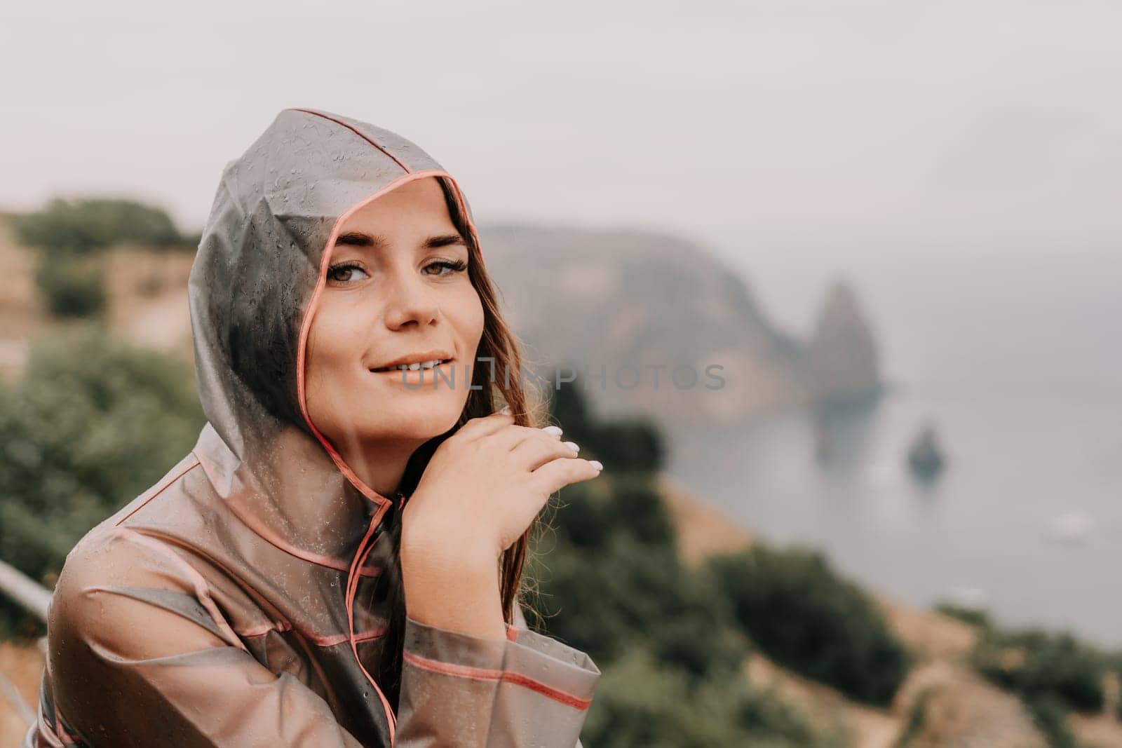 Woman rain park. Happy woman portrait wearing a raincoat with transparent umbrella outdoors on rainy day in park near sea. Girl on the nature on rainy overcast day