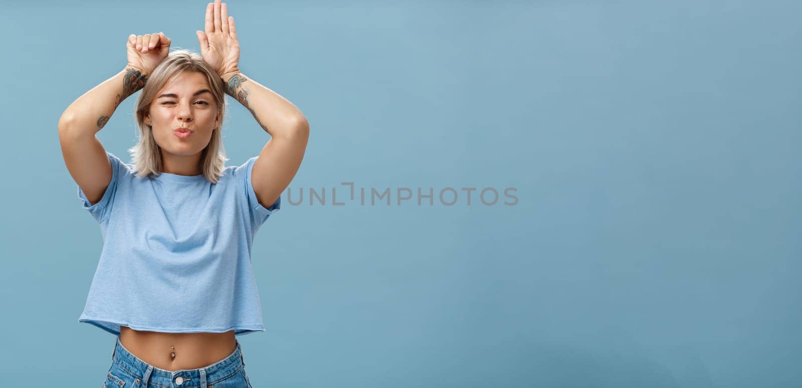 Lifestyle. Flirty attractive and stylish cool girlfriend with tattooed arms and pierced belly winking folding lips in air kiss showing bunny ears with palms on head standing over blue background playful.