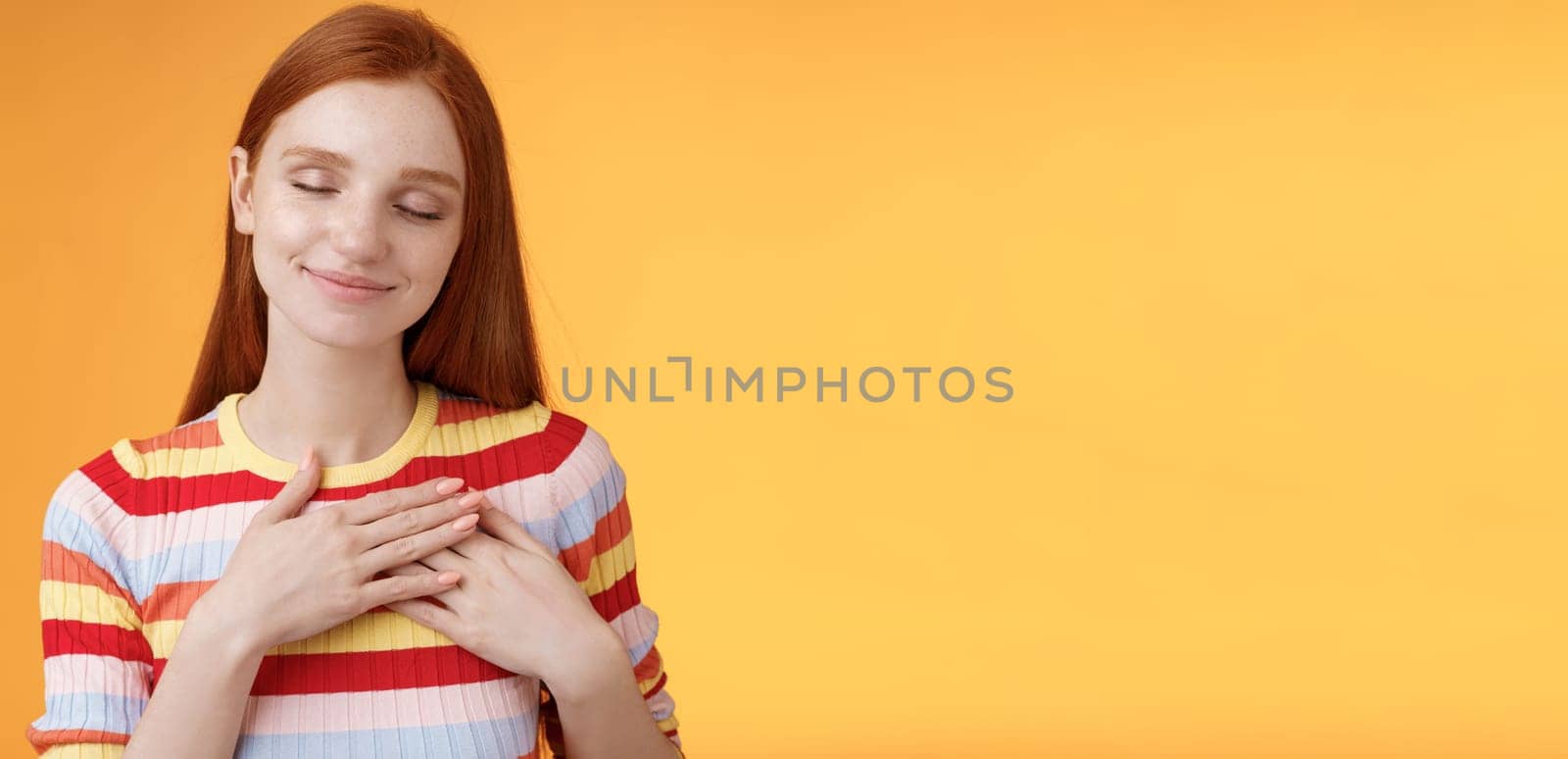Heart keeps warm save dearest memories inside. Smiling tender heartwarming redhead lovely girl touch chest grinning closed eyes recall lovely nice moment standing romantic orange background.