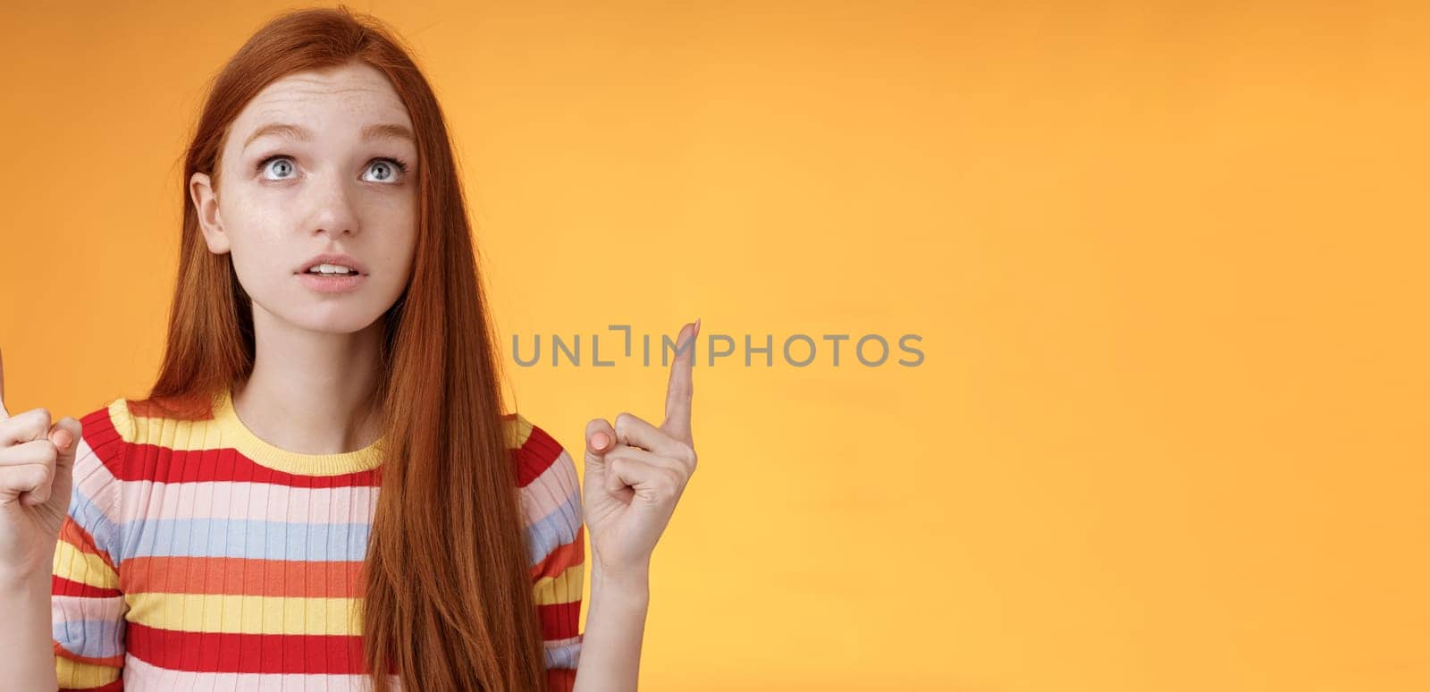 Stunned thrilled young redhead woman peer focused pointing up index fingers upwards look concentrated excited hold breath amused performance standing orange background intrigued and curious.