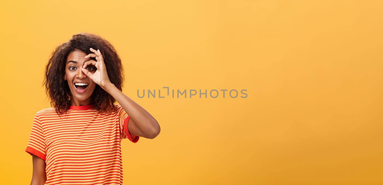 Creative and playful dreamy dark-skinned adult girl with curly hairstyle in striped t-shirt showing circle over eye or okay gesture smiling broadly ready take part in adventures near orange wall. Lifestyle.