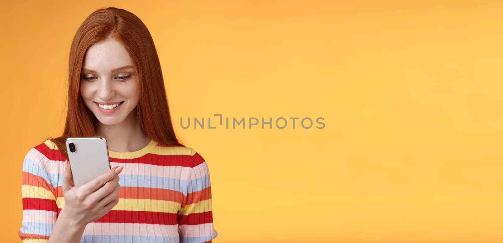 Charming modern redhead girl college student checking message box holding smartphone look happy smiling delighted cellphone display receive hundred likes photo post online, orange background.