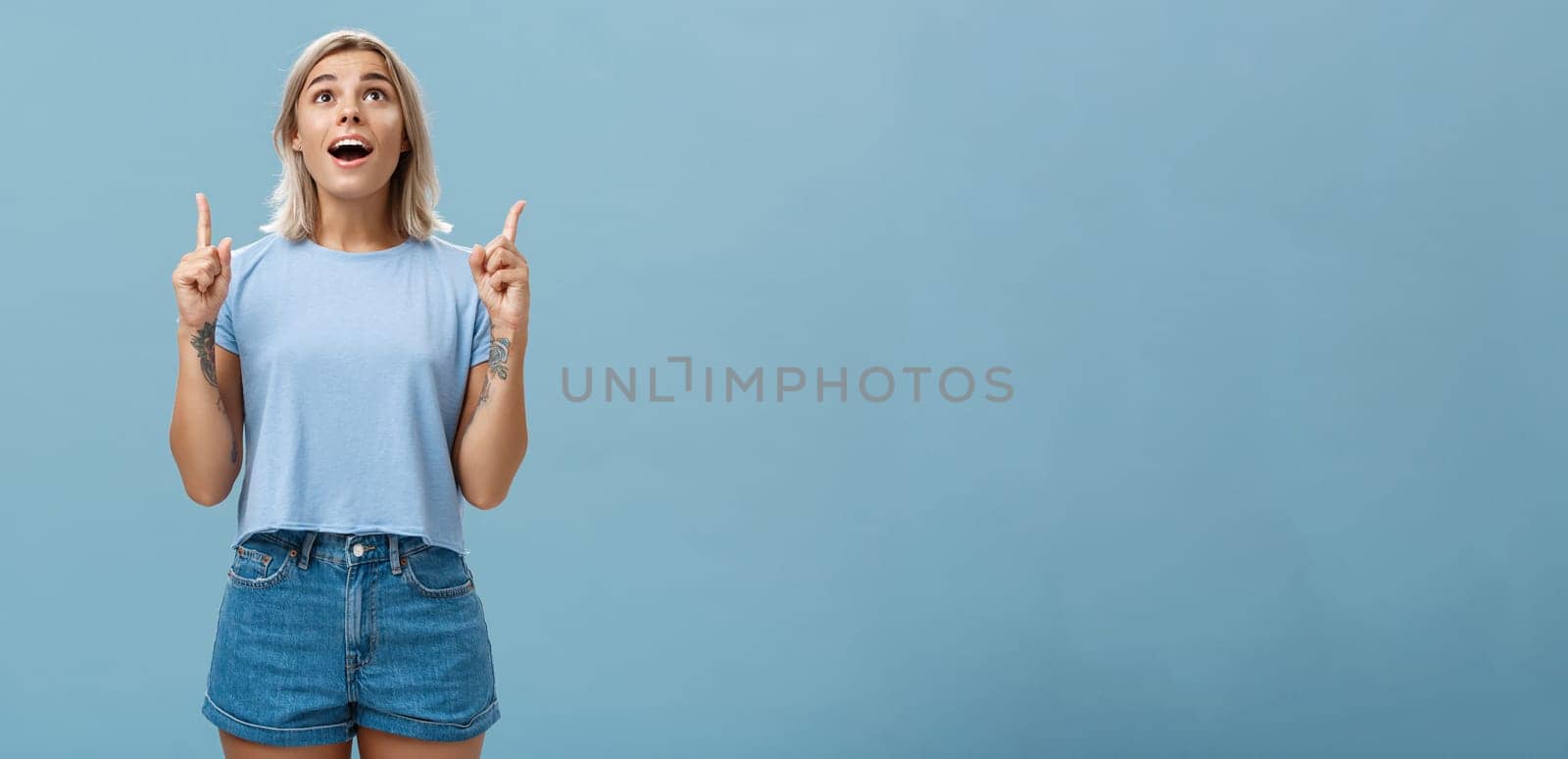 Indoor shot of impressed speechless attractive fair-haired female student in casual t-shirt and denim shorts dropping jaw from amazement pointing and looking up intrigued over blue wall by Benzoix