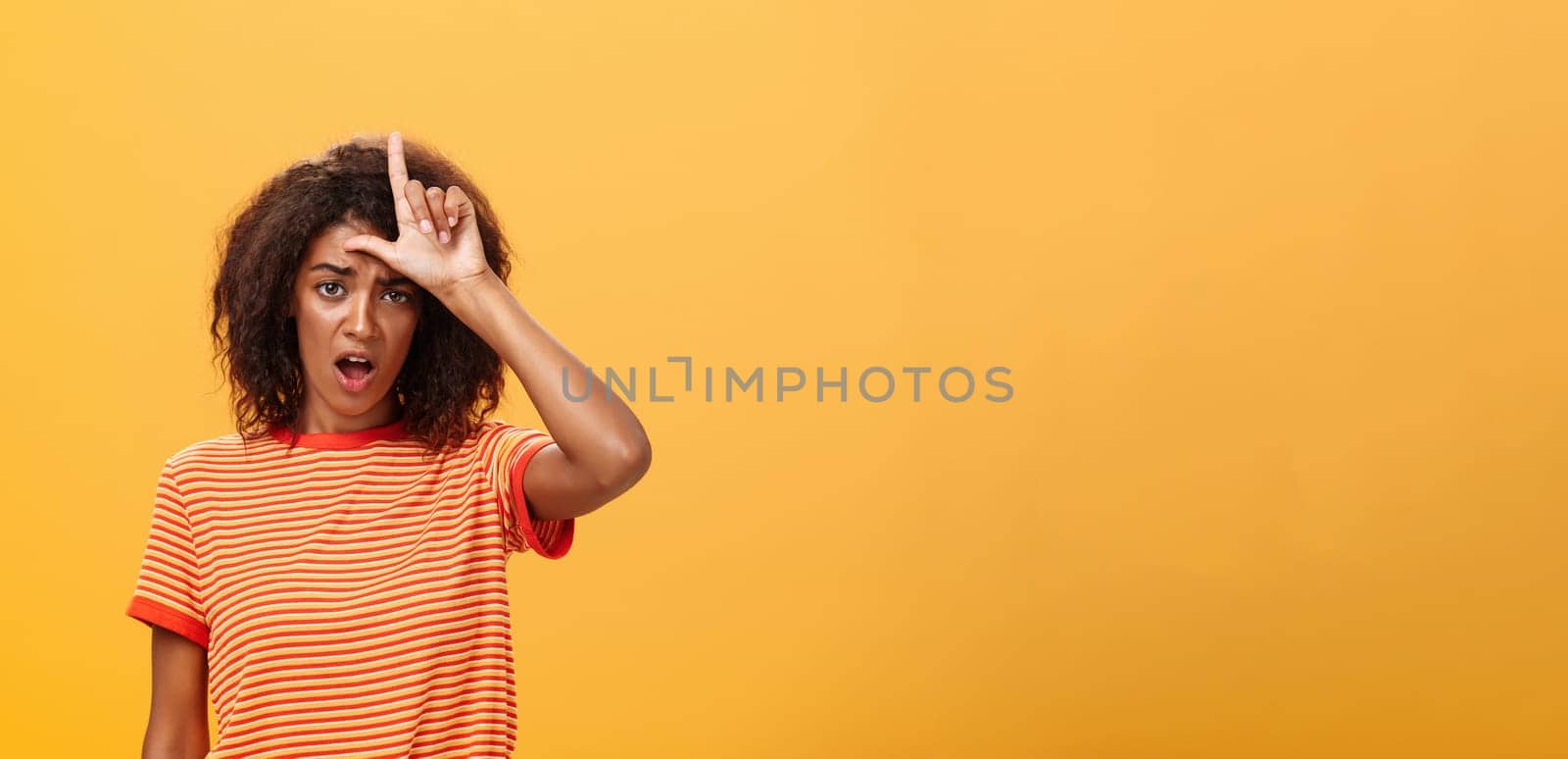 Girl thinks she loser. Portrait of gloomy bothered and displeased african american woman with afro hairstyle showing l word over forehead complaining feeling gloomy and unhappy over orange background.