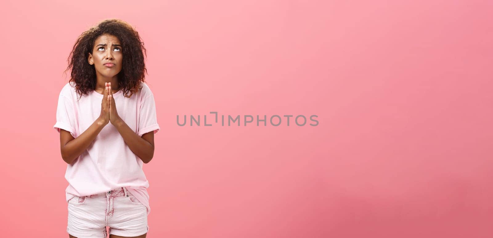 Studio shot of unhappy miserable and hopeless cute african american female holding hands in pray near chest looking up with serious-looking concerned expression making wish to god over pink wall by Benzoix
