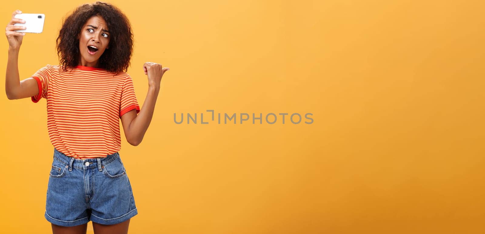 Woman recording video blog pointing at strange object behind her. Portrait of concerned and curious stylish famous internet star holding smartphone talking in cellphone camera over orange background. Technology concept