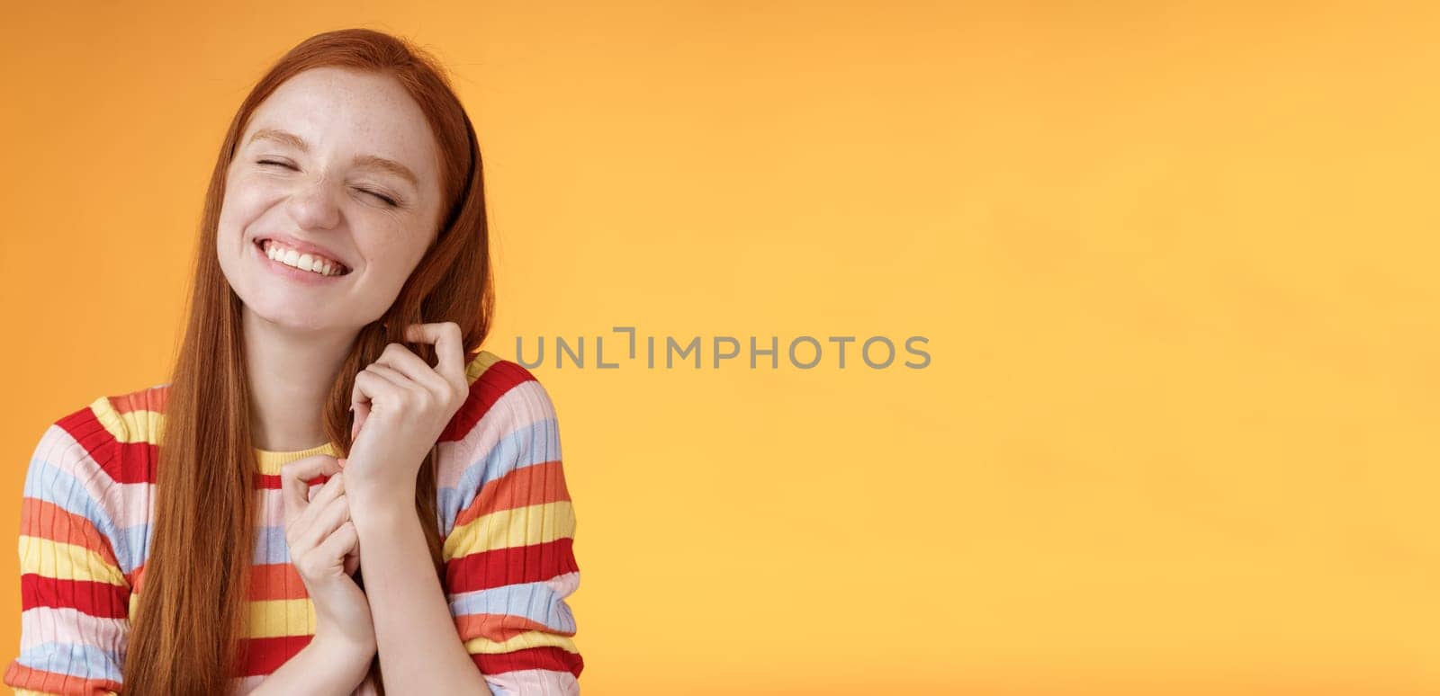 Happy dreamy romantic young tender ginger girl fantasizing creating love story imagination smiling broadly delighted close eyes touching hair strands recalling nice memory, standing orange background.