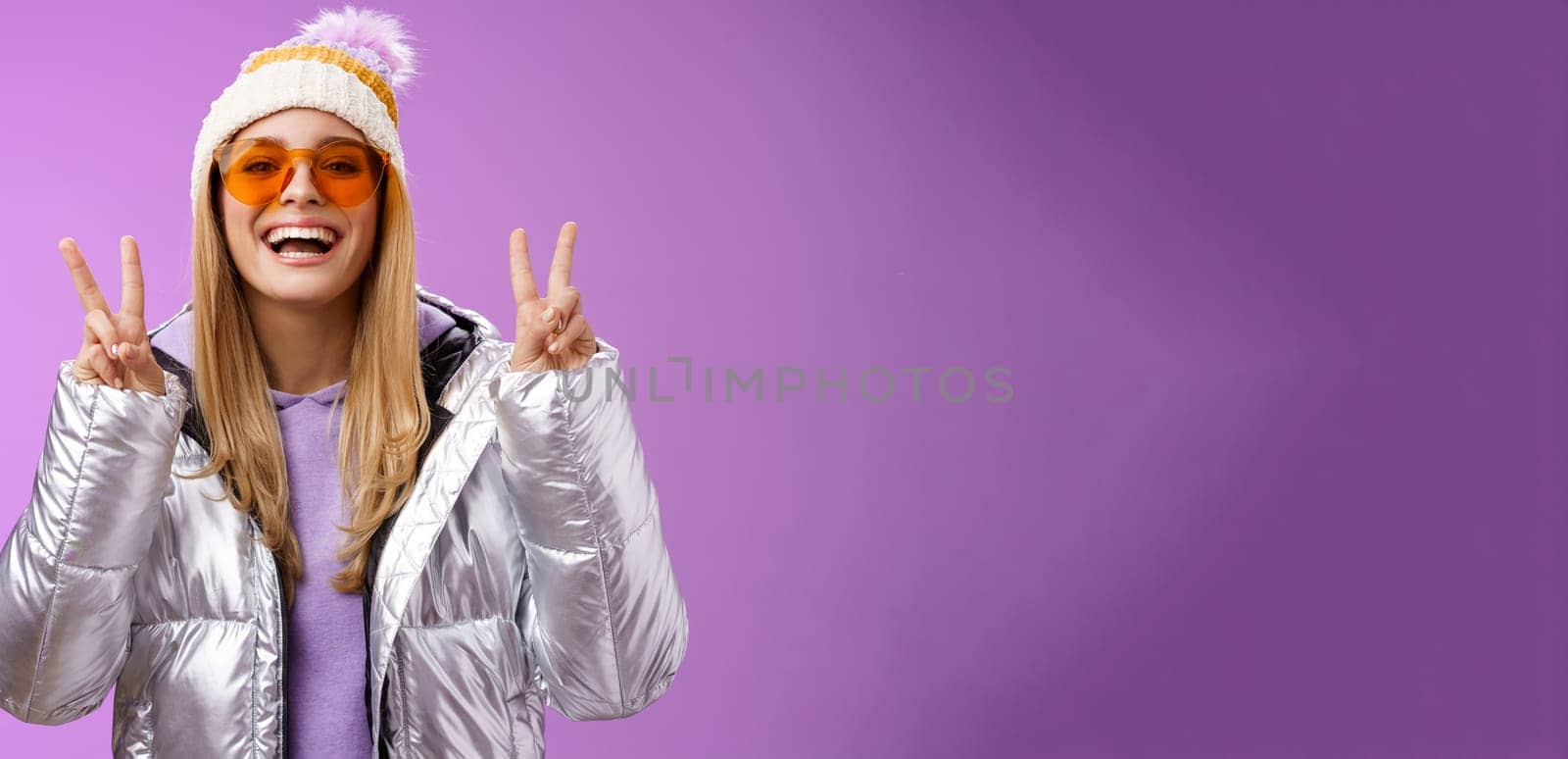 Friendly lucky optimistic beautiful blond woman in sunglasses silver stylish jacket winter hat show peace victory gesture enjoying snowy vacation get outside hotel enjoy skiing, purple background.