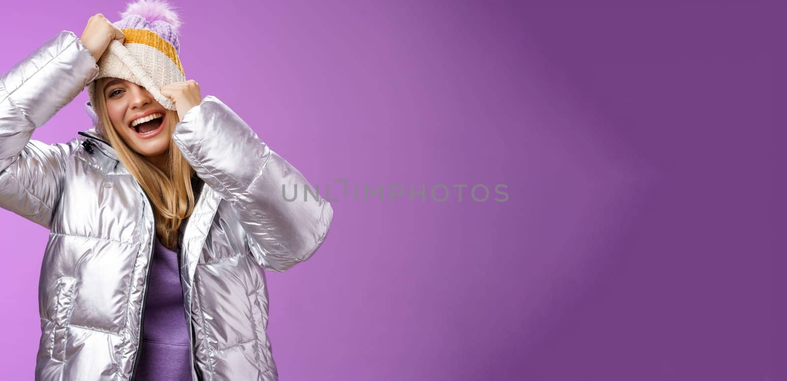 Amused carefree happy smiling playful attractive woman having fun laughing joyfully pulling hat face hiding peeking one eye camera grinning chuckling fool around have fun, purple background.