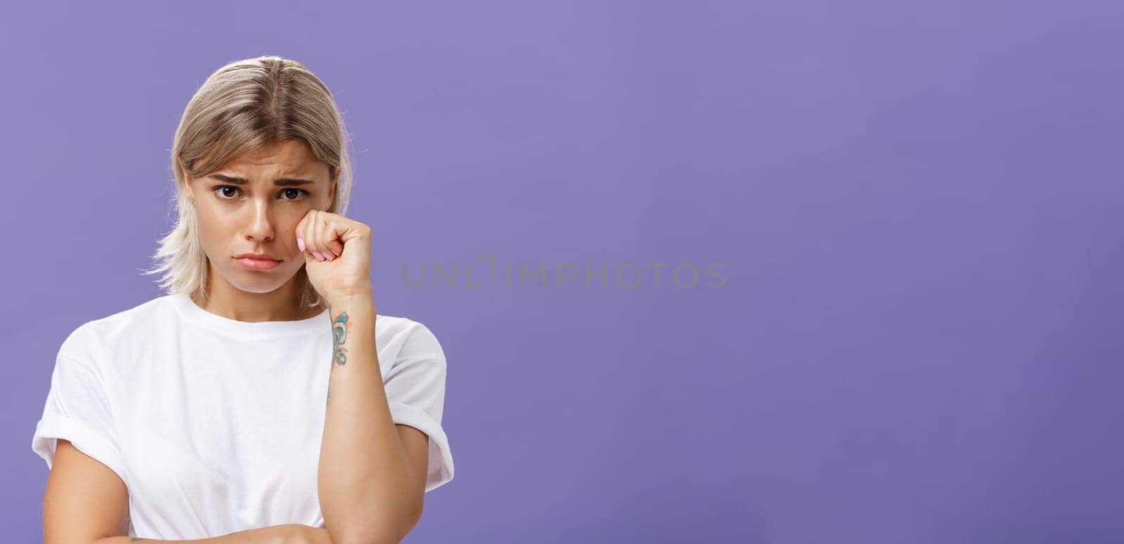 Studio shot of offended sad and timid silly woman with blond hairstyle frowning looking from under forehead holding fist near eye as if whiping teardrop being upset over purple background by Benzoix