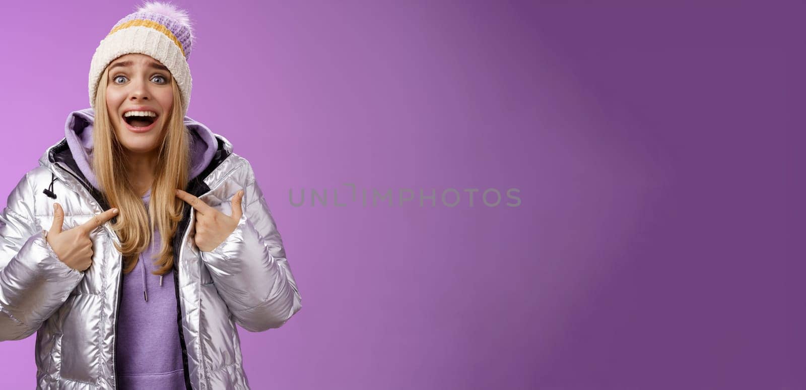 Surprised happy smiling joyful attractive woman pointing herself satisfied picked was chosen winner standing excited in silver shiny jacket winter hat cannot believe own luck, purple background.