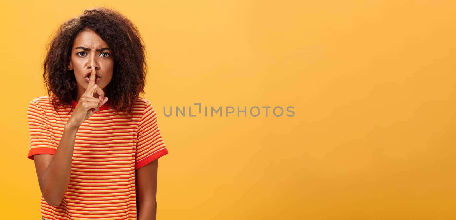 Shh do not disturb. Portrait of serious-looking bossy and displeased cute african american girl with afro hairstyle frowning shushing at camera with index finger over mouth forbidding speak loud.