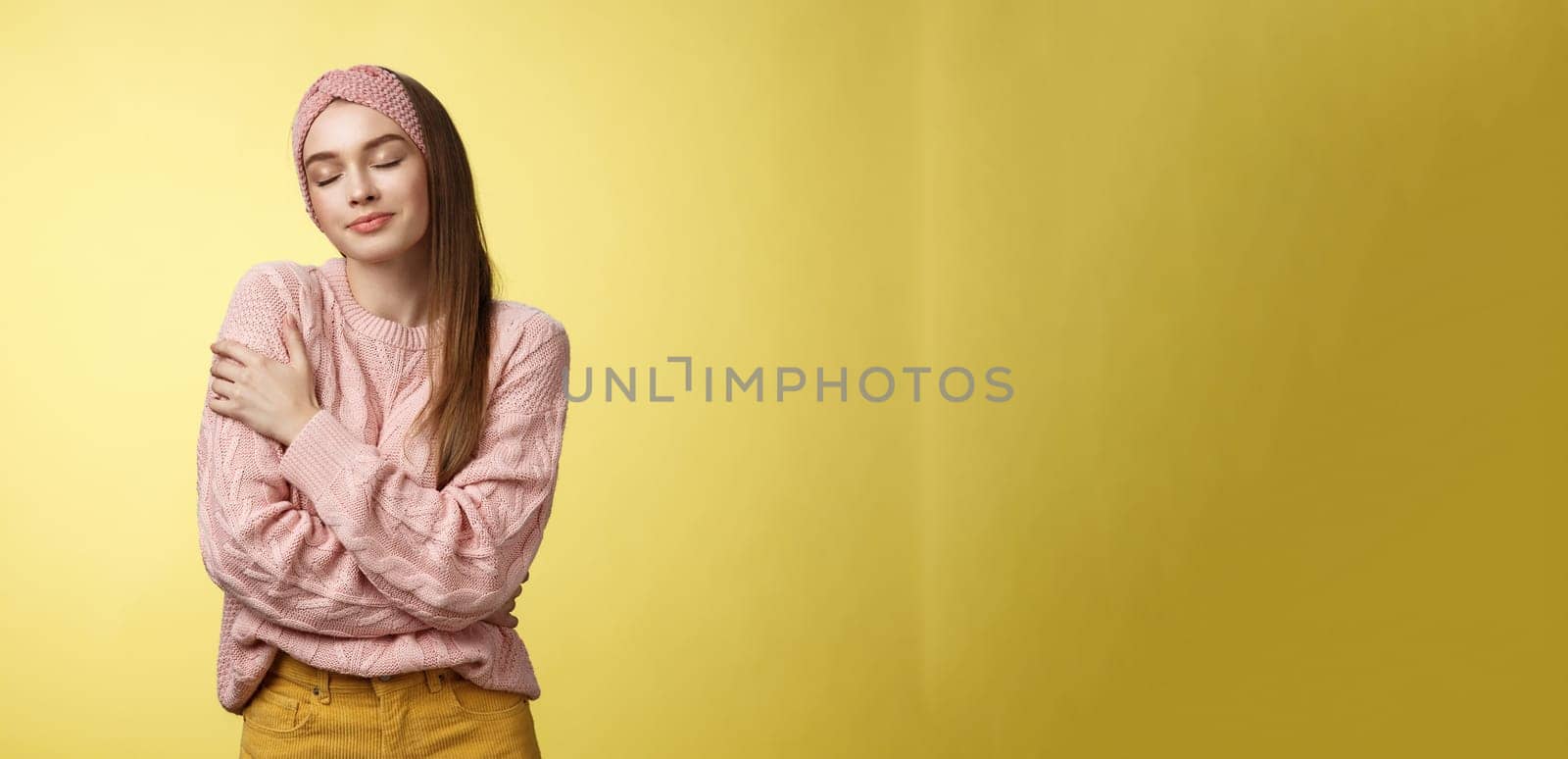 Sweet glamour young girl wearing warm comfy sweater embracing herself crossing arms over body in hug, smiling soft and kind close eyes, recalling lovely days, tender memories over yellow wall.