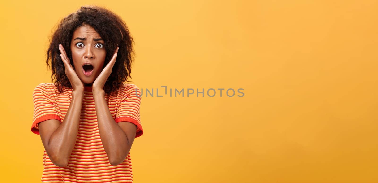 Waist-up shot of shocked concerned panicking african-american woman with afro hairstyle in trendy t-shirt gasping holding palms on face from surprise posing troubled against orange wall. Lifestyle.