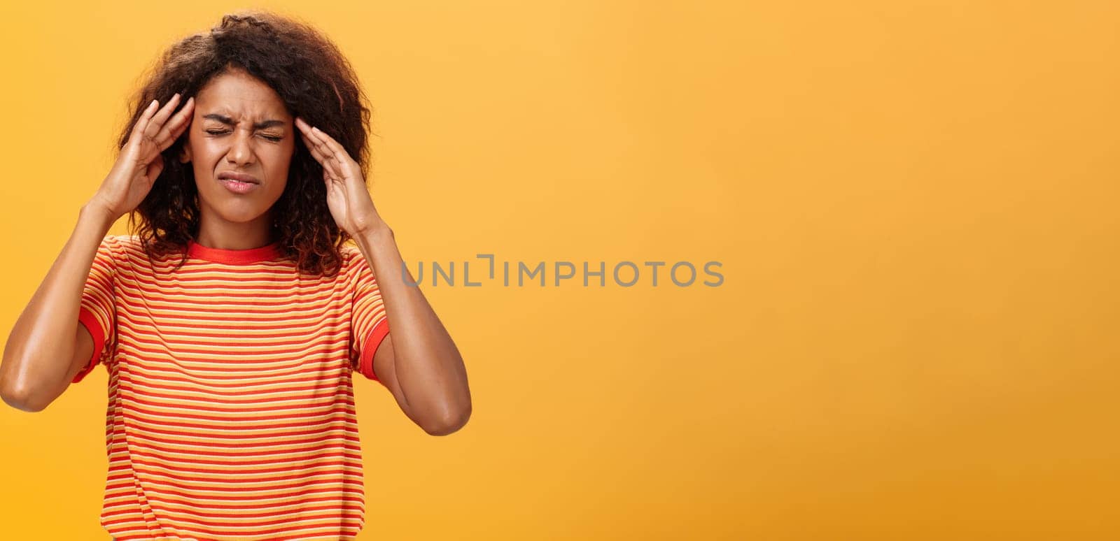 Woman cannot recall important information looking intense trying concetrate under pressure feeling tensed touching temples closing eyes while thinking hard posing against orange background.