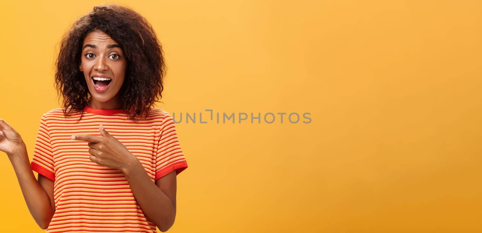 Portrait of amazed excited charismatic dark-skinned young pretty girl with afro hairstyle in trendy striped t-shirt pointing left delighted and fascinated posing against orange background. Lifestyle.
