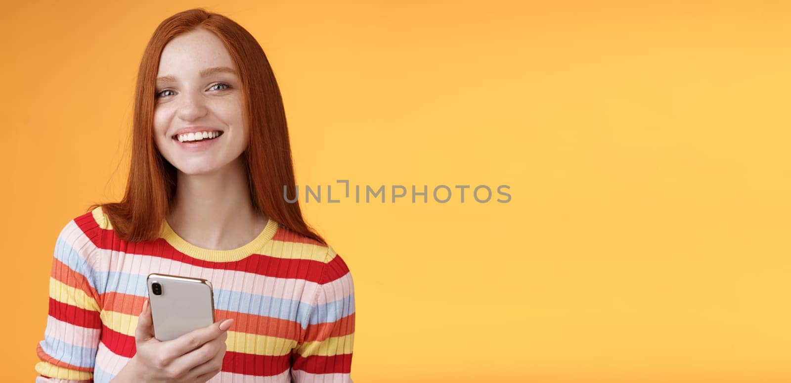 Friendly enthusiastic young redhead girl blue eyes using smartphone turn camera answer smiling broadly telling who sent message standing delighted orange background messaging, texting boyfriend.
