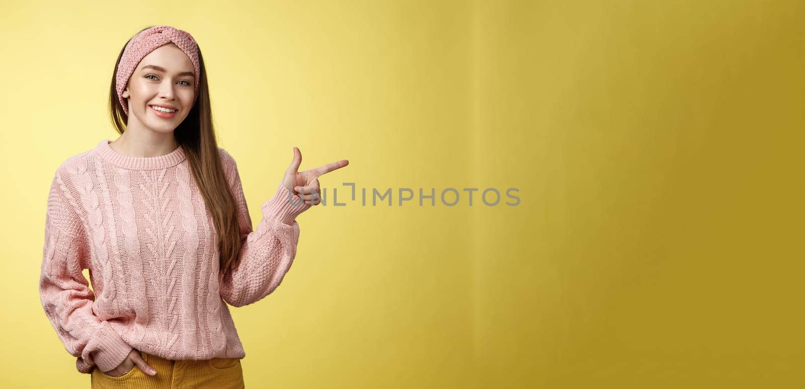 Lifestyle, people, pointing concept. Attractive young girl with long straight hair wearing sweater pointing aside using fore finger, indicating copy space smiling cheerful, positive over yellow wall by Benzoix