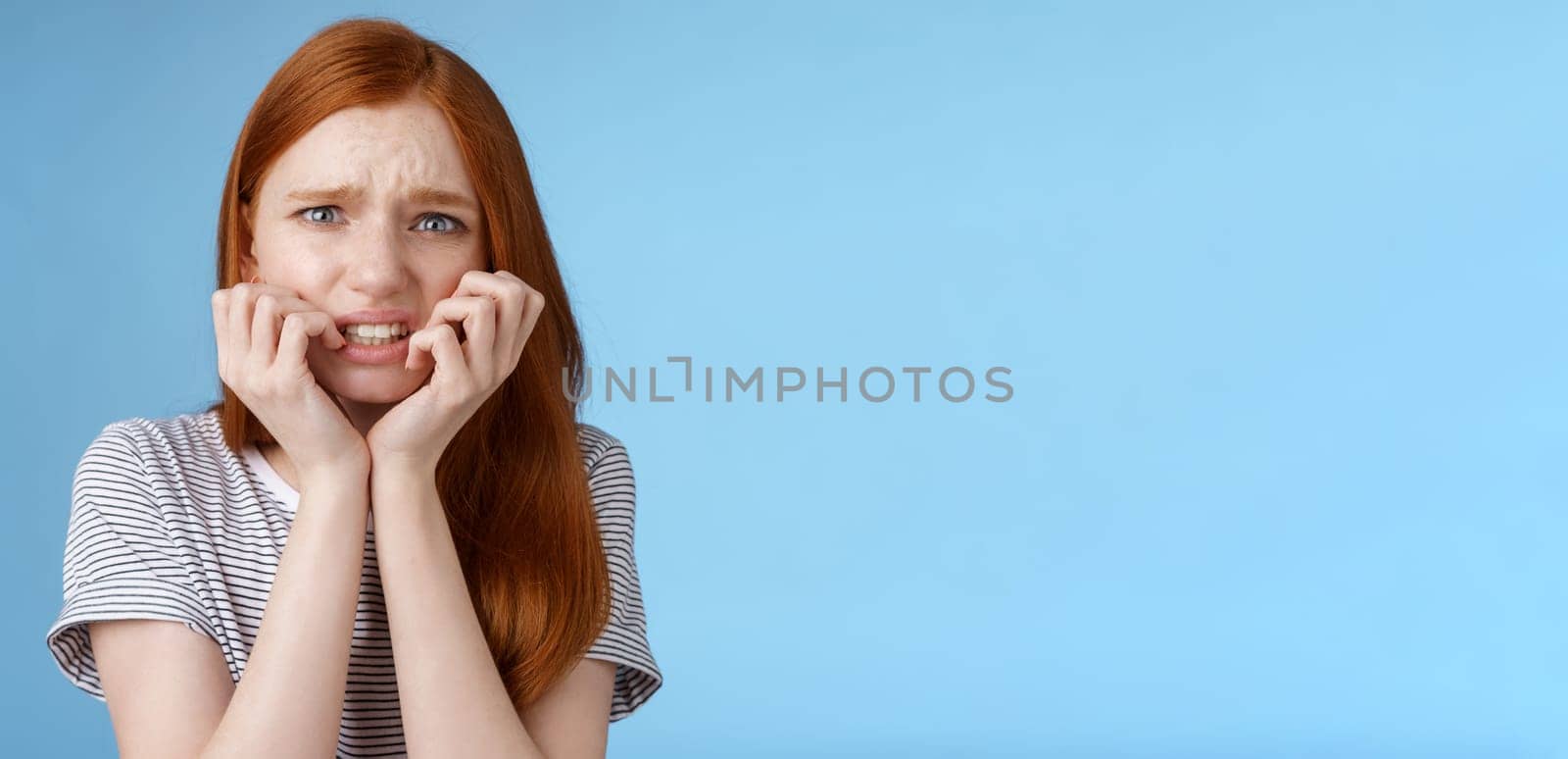 Worried uncomfortable scared young panicking redhead young girl feeling pressure distressed frowning squinting frightened biting fingernails trembling fear, standing blue background.