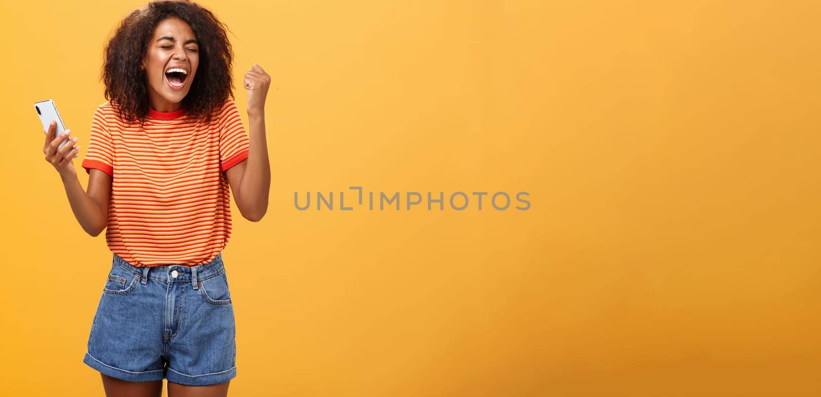 Portrait of ambitious happy young african american girl yelling from happiness and triumph clenching fist in joy and celebration feeling excited and relieved holding smartphone over orange wall. Lifestyle.