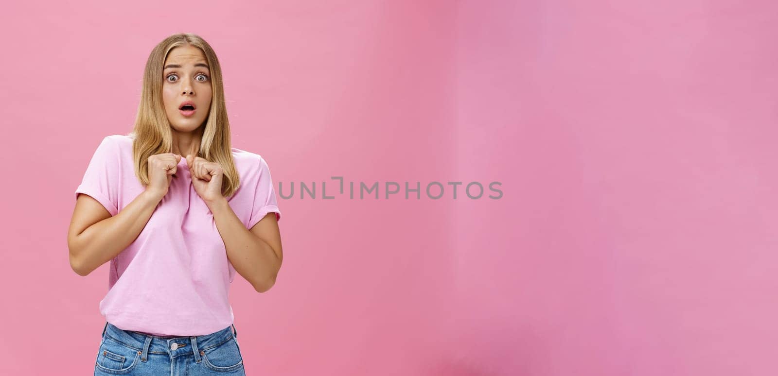 Woman gasping from fright being scared by someone pressing clenched hands to chest bending backwards looking worried and terrified at camera being shocked with unexpected bang over pink wall by Benzoix