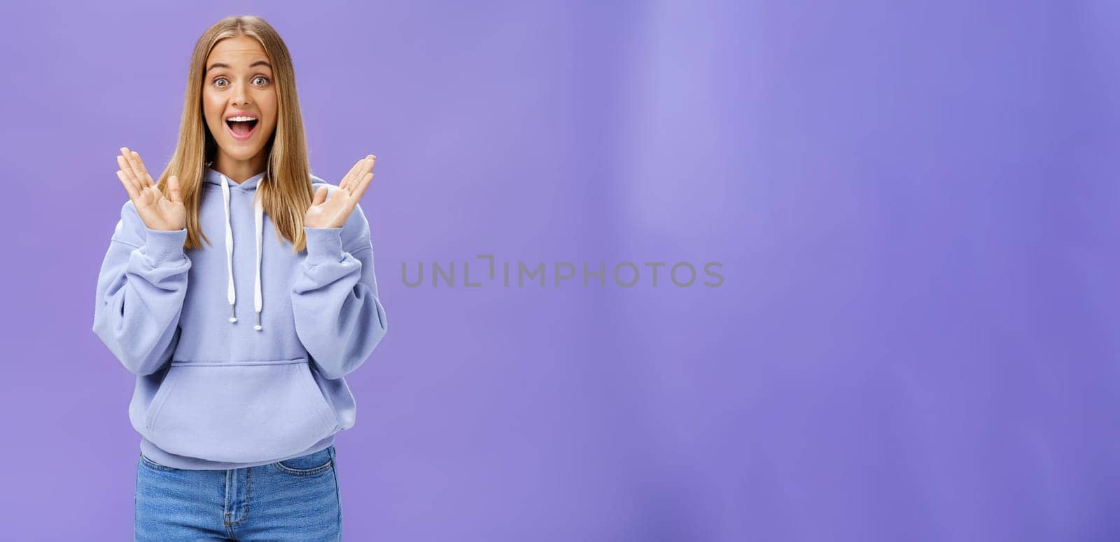 Portrait of amused excited and entertained happy charming woman with fair hair in trendy hoodie clasping hands from delight and surprise smiling broadly joyfully posing against purple background by Benzoix