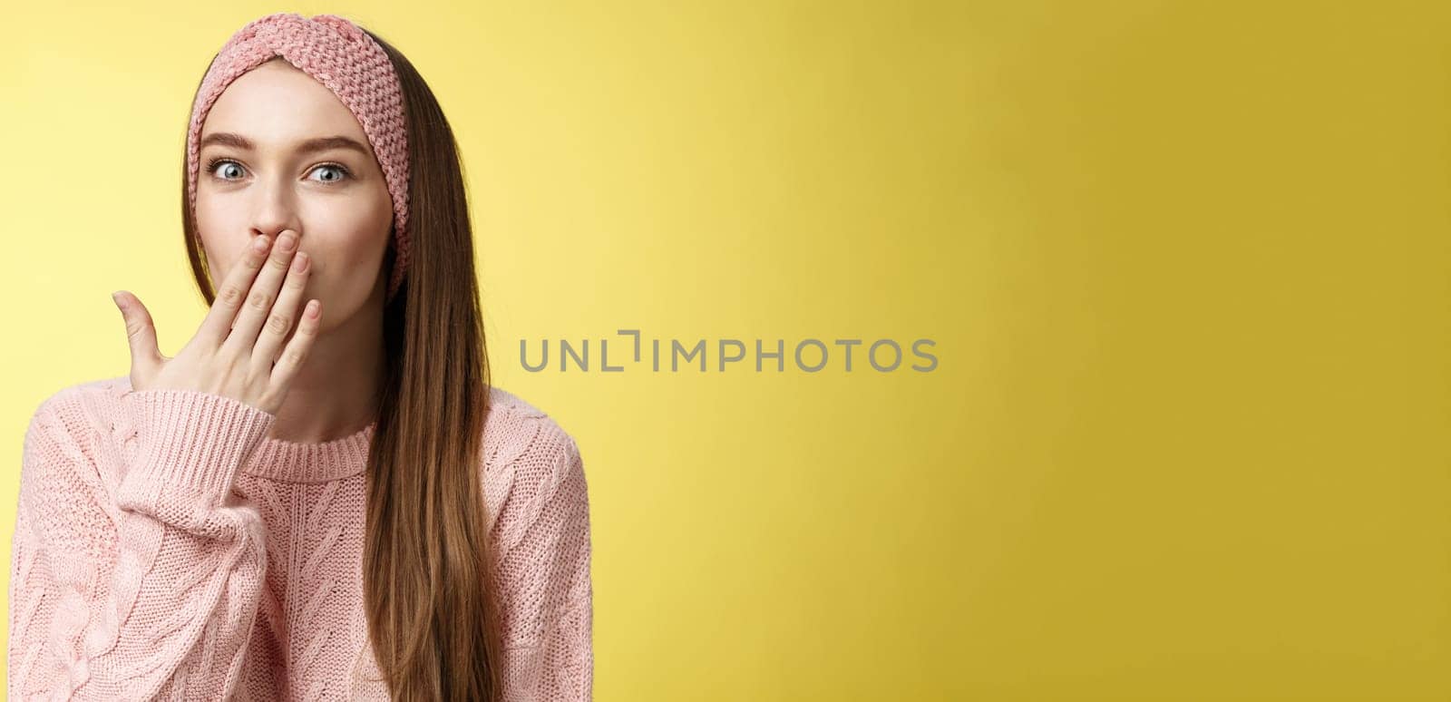 Talkative young cute office worker wearing sweater, knitted headband holding palm on mouth surprised and amazed, learning interesting shocking rumor, gossiping entertained over yellow wall.