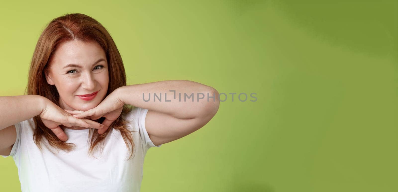 Aging, cosmetology, wellbeing concept. Happy self-assured redhead woman hold hands under jawline smiling showing facial blemished self-accepting wrinkles applying skincare product green background.