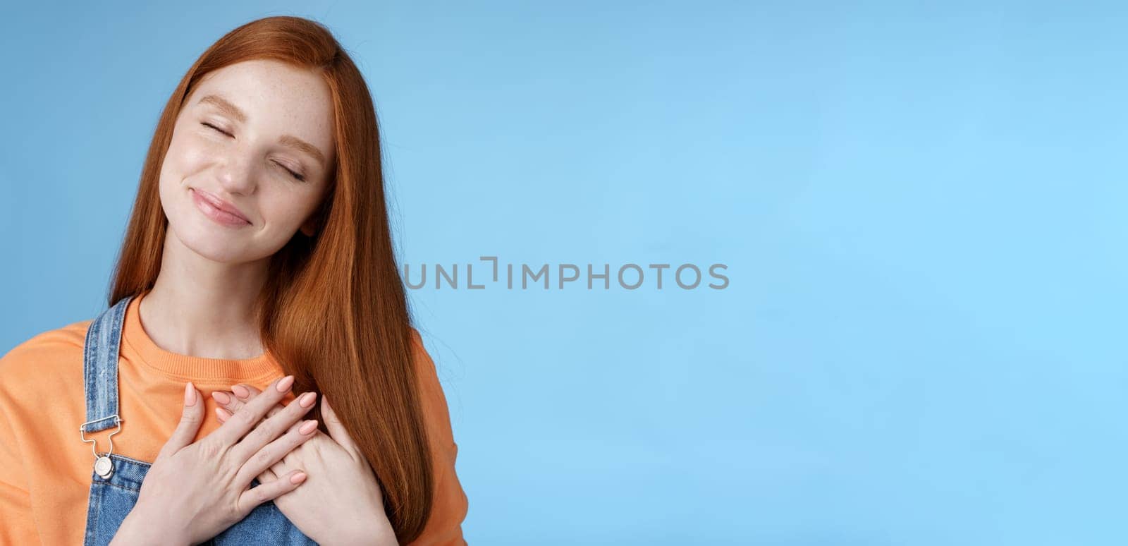 Dreamy touched romantic ginger girlfriend close eyes recalling heartwarming romance touch heart palms pressed chest smiling tenderly feel love care sympathy express affection, blue background by Benzoix