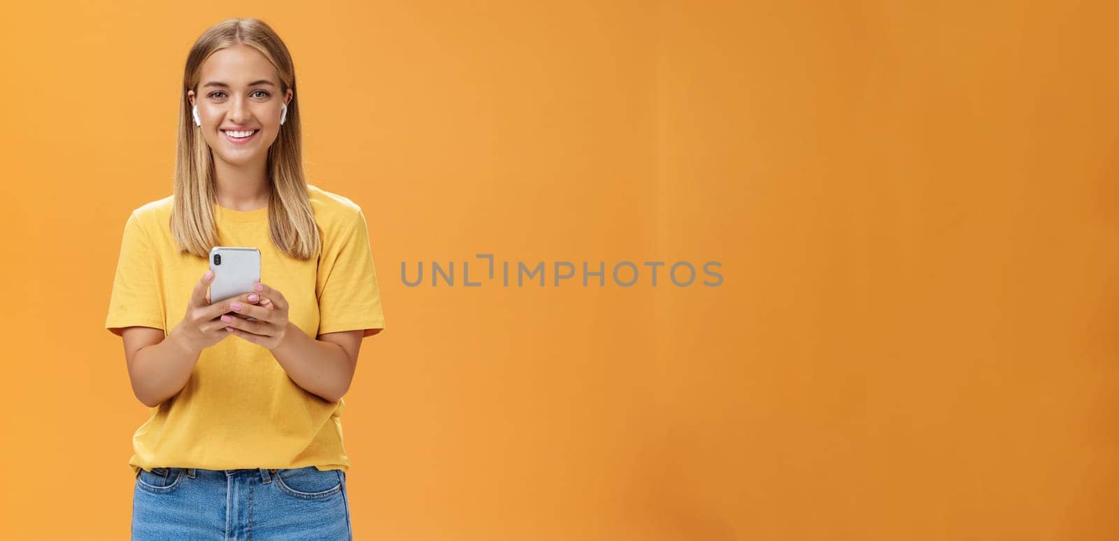 Portrait of friendly charismatic and outgoing good-looking young female in yellow t-shirt holding smartphone wearing wireless earphones enjoying listening music on way to fitness gym. Technology and lifestyle concept