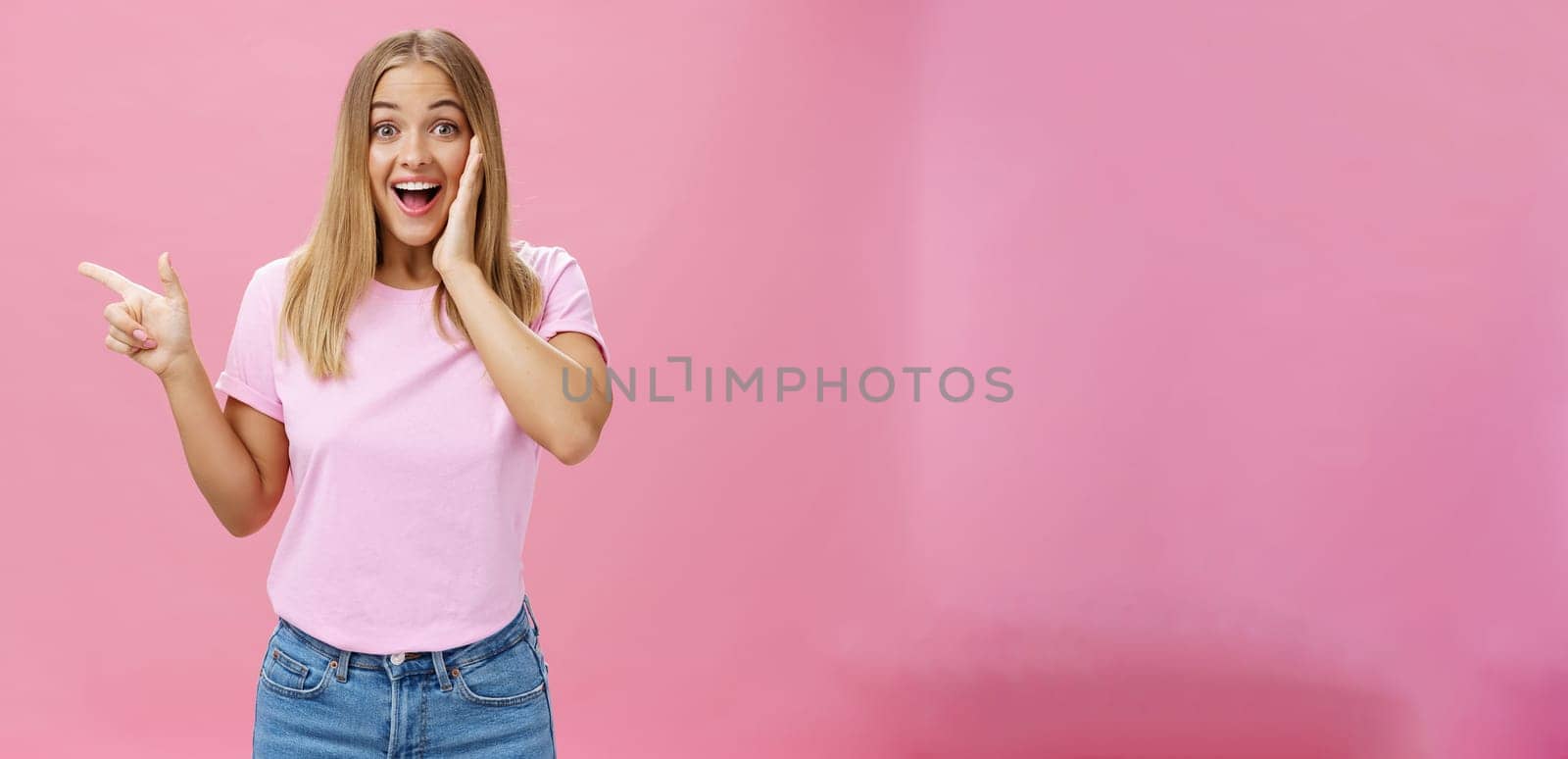 Portrait of surprised enthusiastic and charismatic tanned cute woman with fair straight hair touching cheek from joy and amazement pointing left delighted and amused over pink background by Benzoix