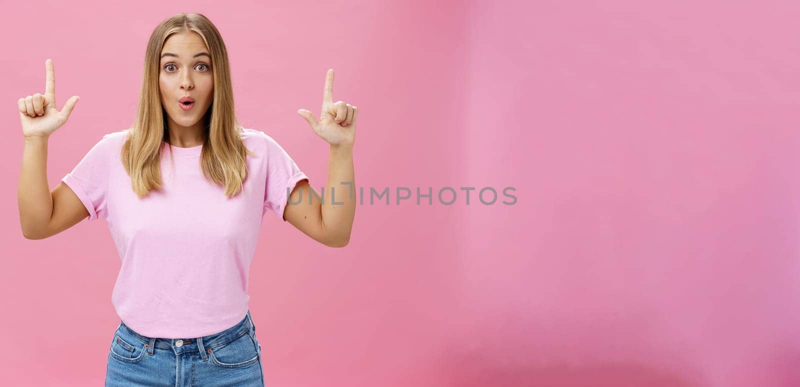 Amused and impressed attractive common european woman with fair hair and tanned skin raising index fingers pointing up amazed folding lips from interest and astonishment over pink background. Copy space