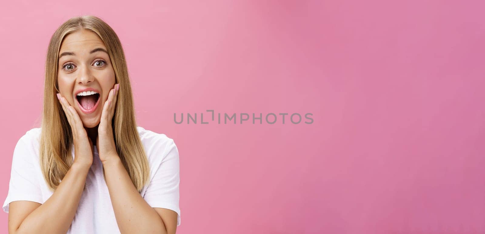 Portrait of amused delighted friendly and attractive european woman with fair hair touching cheeks from amusement and surprise smiling broadly gazing with admiration at camera over pink background by Benzoix