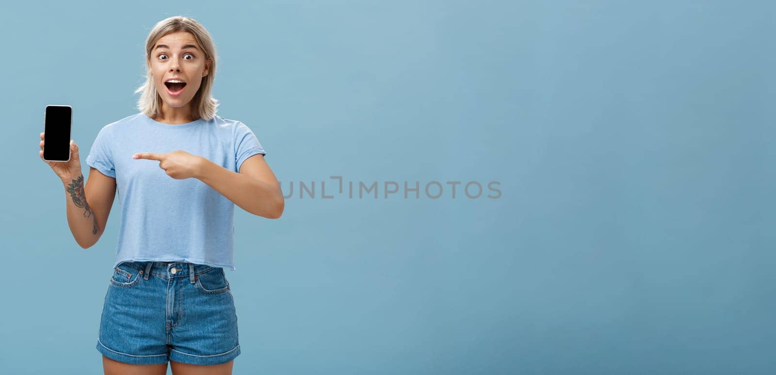 Studio shot of amazed and impressed charming european blonde in casual clothes gasping charmed at camera while showing smartphone screen pointing at device with finger over blue background by Benzoix