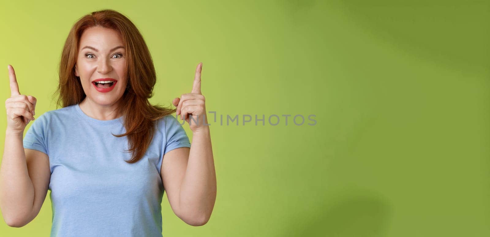 Impressed enthusiastic happy cheerful middle-aged mature woman red hair smiling astonished look admiration joy pointing up raised index fingers introduce impressive stunning offer green background by Benzoix